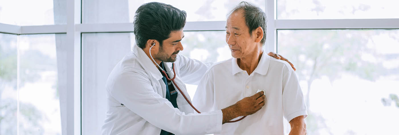 A male physician wearing a white coat uses his stethoscope to listen to the heart of his patient, an older Asian man wearing a white polo shirt. They are seated in front of a large window with green trees outside. 