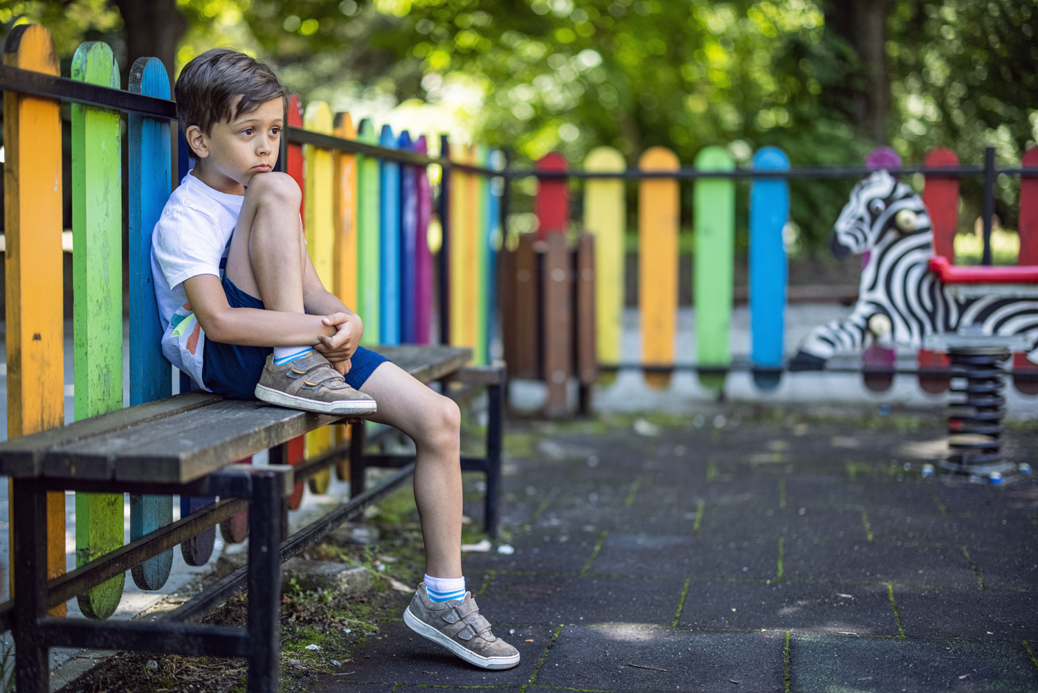 A young boy sits against a fence in a playground, appearing concerned.