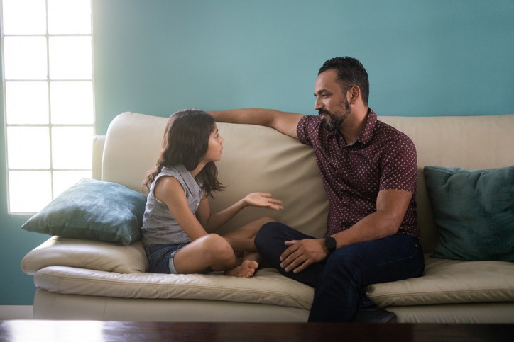 A father and his daughter sit on a couch having a conversation.