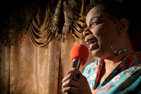 Debra Faulk, a Black woman, holds a microphone as she stands onstage, with heavy gold curtains behind her, performing a standup comedy set.