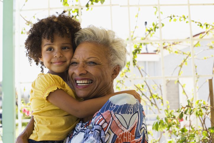 A Black grandmother holds her grandchild as both smile, looking left out of the frame