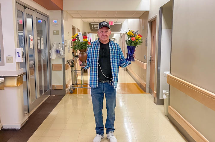 A middle-aged man stands in a hospital hallway holding two colorful flower bouquets in vases, one in each hand.