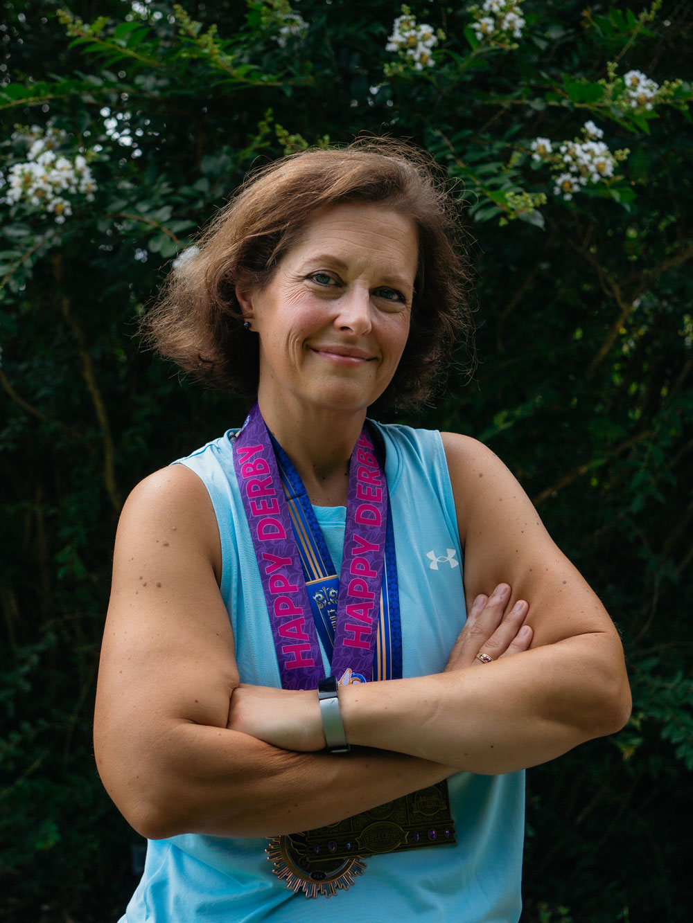 Jeanne Hartman smiles at a camera with her arms crossed over her chest, wearing several medals from running competitions 