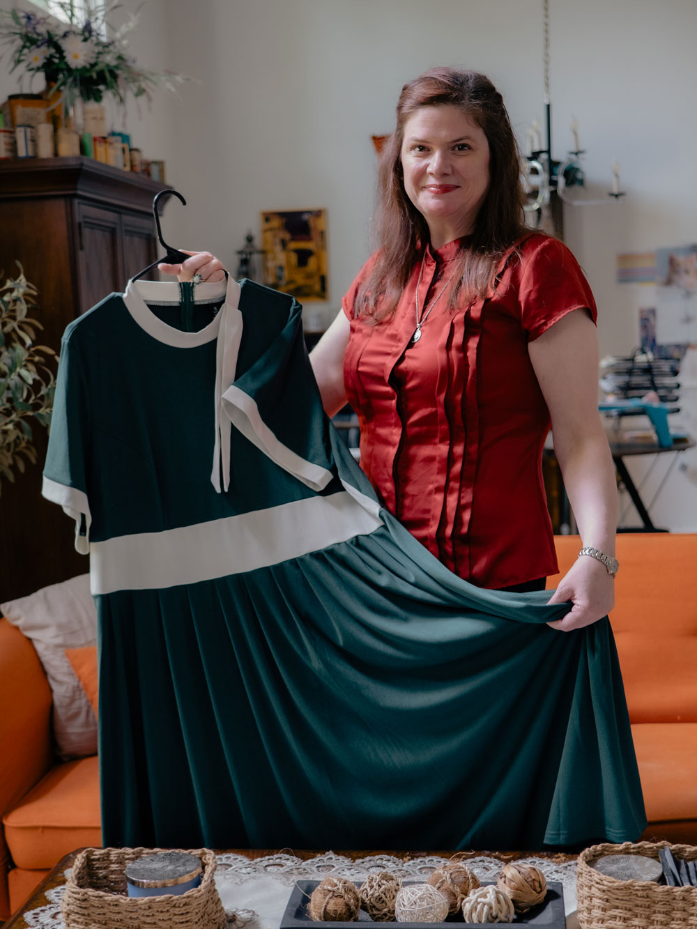 Nicola Allen, wearing a red shirt and a necklace, smiles toward camera while holding a black dress. She's standing in a room full of sewing materials.