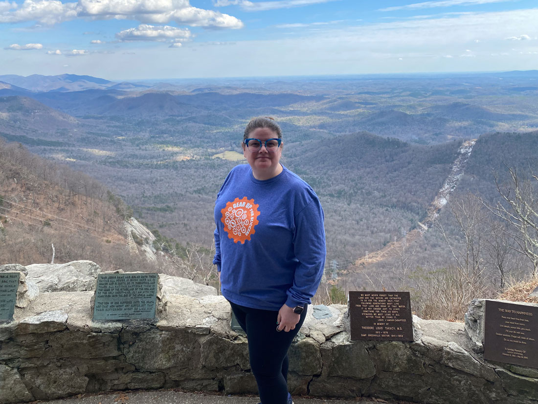 Nicola Allen, wearing a blue long-sleeved tee shirt with a green gear that says "Gear Up For Success," standing for a photo in front of a mountain overlook area.