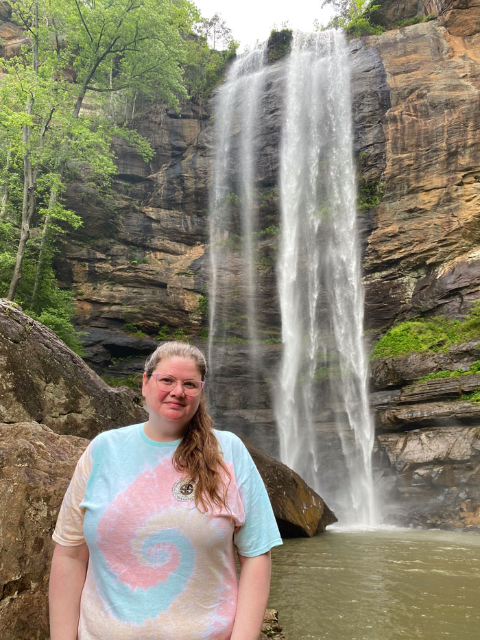 Nicola Allen is close to the foreground of a photo, wearing a tie-dye shirt. She's standing in front of a small pond being filled by a small waterfall.