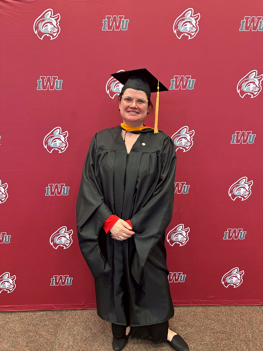 Nicola Allen dressed in graduation gown. She's smiling gleefully in front of a banner that indicates she is at Indiana Wesleyan University.