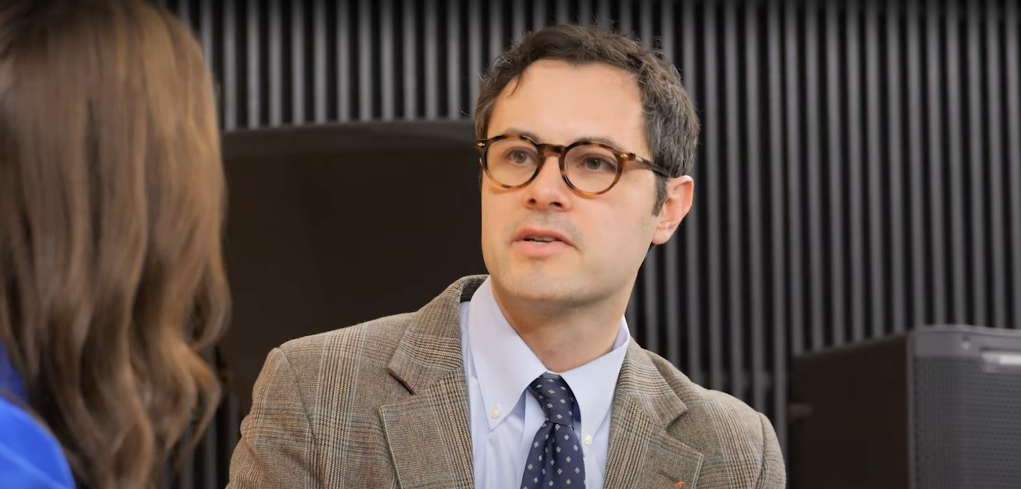 Dr. David Neil Toupin looks at a woman who's partially off-screen. He's seated and wearing a suit.
