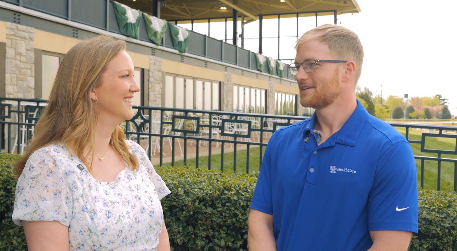 A woman in a dress patterned with blue flowers, left, speaks to a man dressed in a blue polo with a UK HealthCare logo. They are present at Keeneland Race Course.