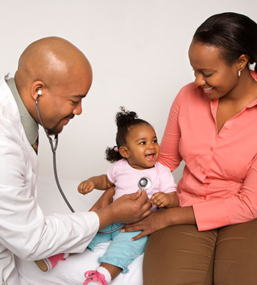 A mother smiles as a male doctor wearing a white coat and stethoscope listens to her toddler daughter's chest.