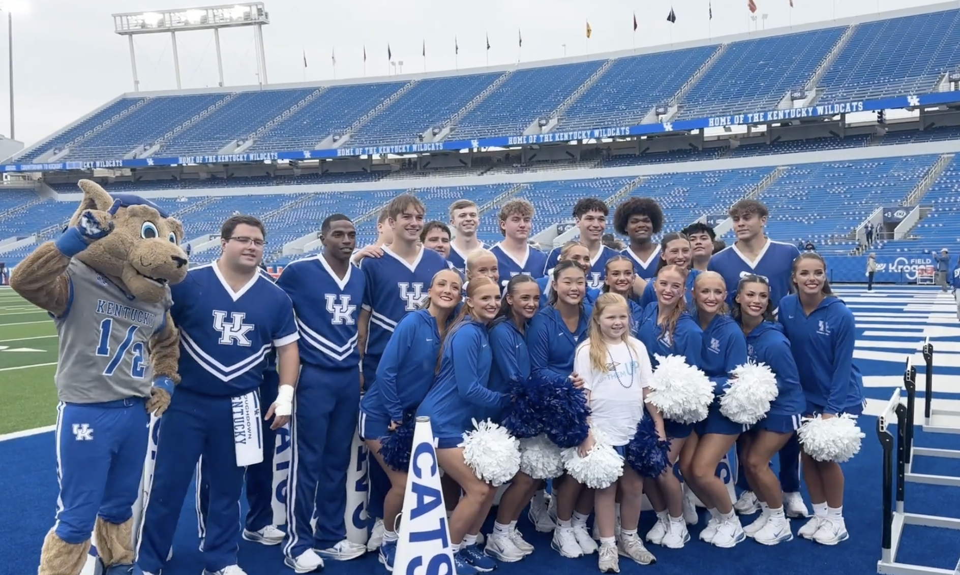 A group of cheerleaders, along with the University of Kentucky mascot, gather around a young girl in the end zone of Kroger Field, a large football stadium