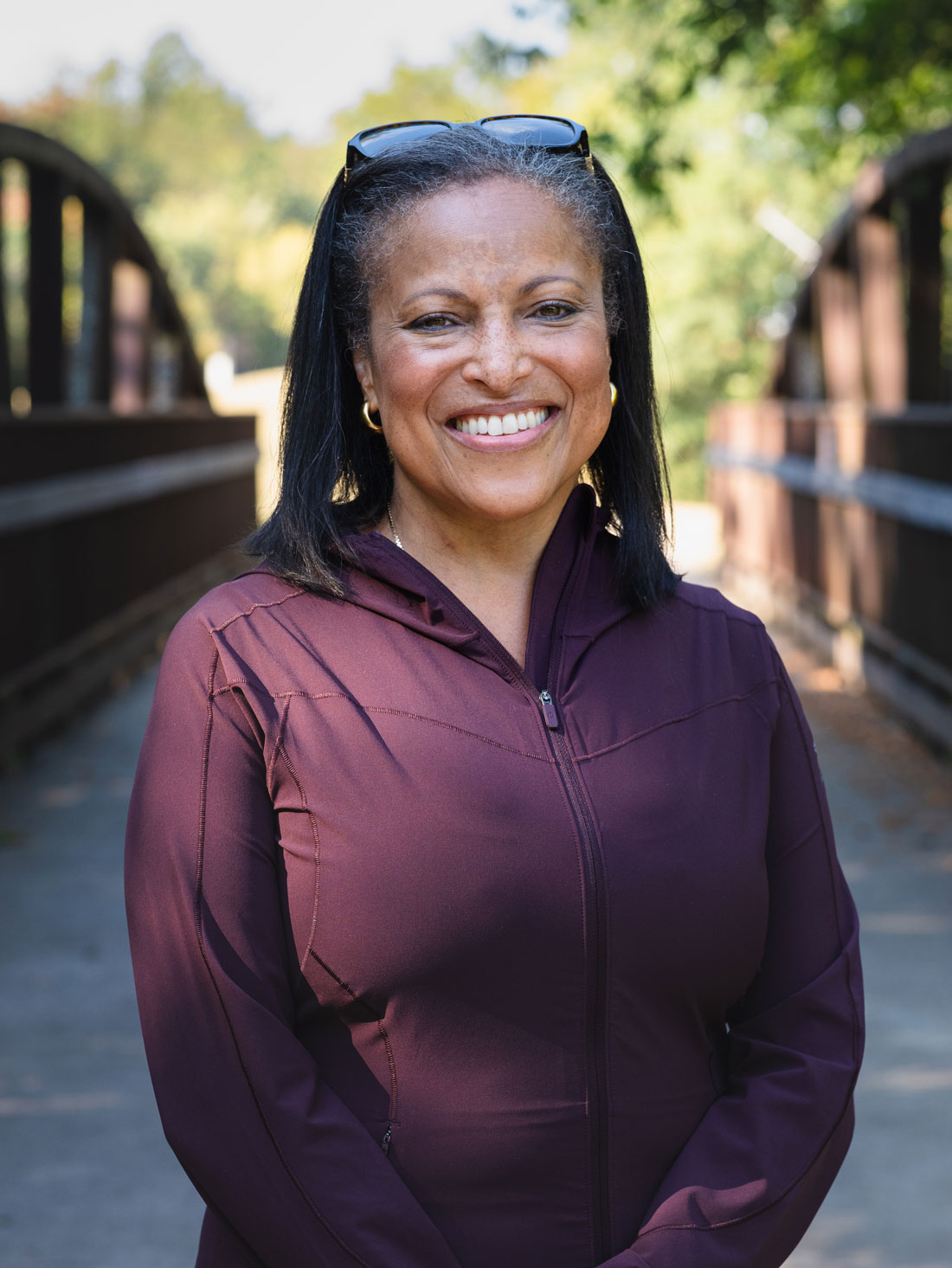 Mid-shot portrait of Maria Braman, who smiles toward the camera while standing on a wooden bridge. She's wearing a black quarter zip hoodie.