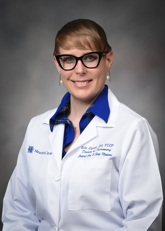 Dr. Erin Camac poses against a gray background wearing a white medical coat with her name written on it.