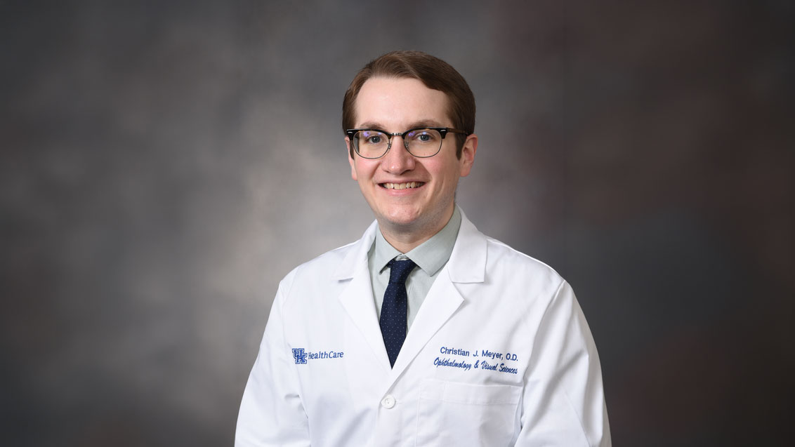 Dr. Christian Meyer smiles while posed against a gray background; the image has been slightly altered in Photoshop to extend the background for widescreen usage