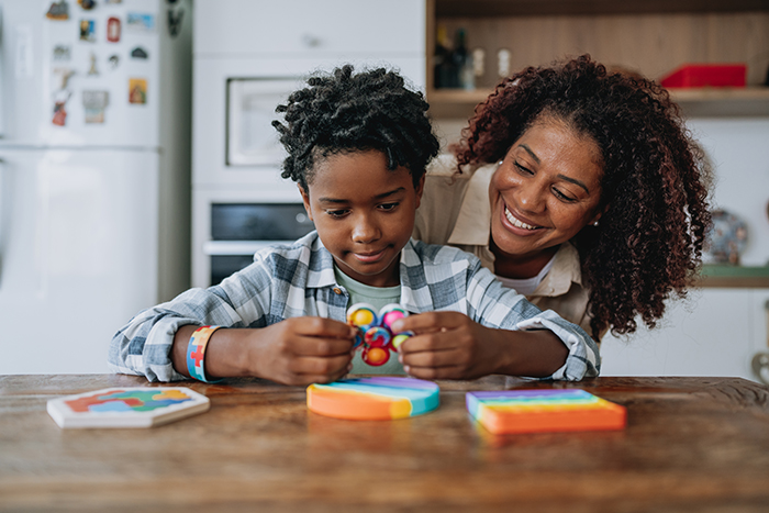 A child sits at a wooden table holding a small, colorful fidget toy, with several brightly colored puzzle or sensory toys arranged on the table. Another person leans in nearby, observing the activity. A kitchen setting with appliances and shelves is visible in the background.