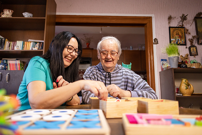 Two people are seated at a table inside a home, working together with wooden puzzle or matching game pieces spread across the surface. Shelving with books and decorative items lines the background, and additional game boards and colorful pieces are visible in the foreground.