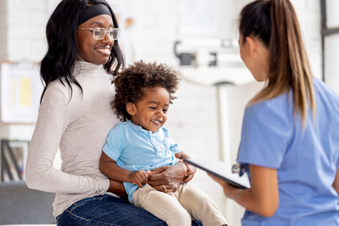 A caregiver holds a toddler during a clinical visit while a healthcare provider reviews information on a tablet.