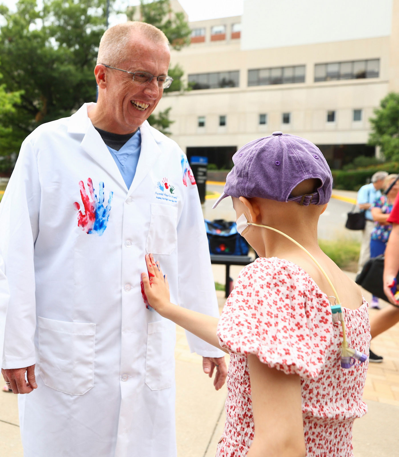 Scottie Day smiling, wearing a coat with a child's handprint painted on it, as a child with an IV in their face sticks a hand to his stomach