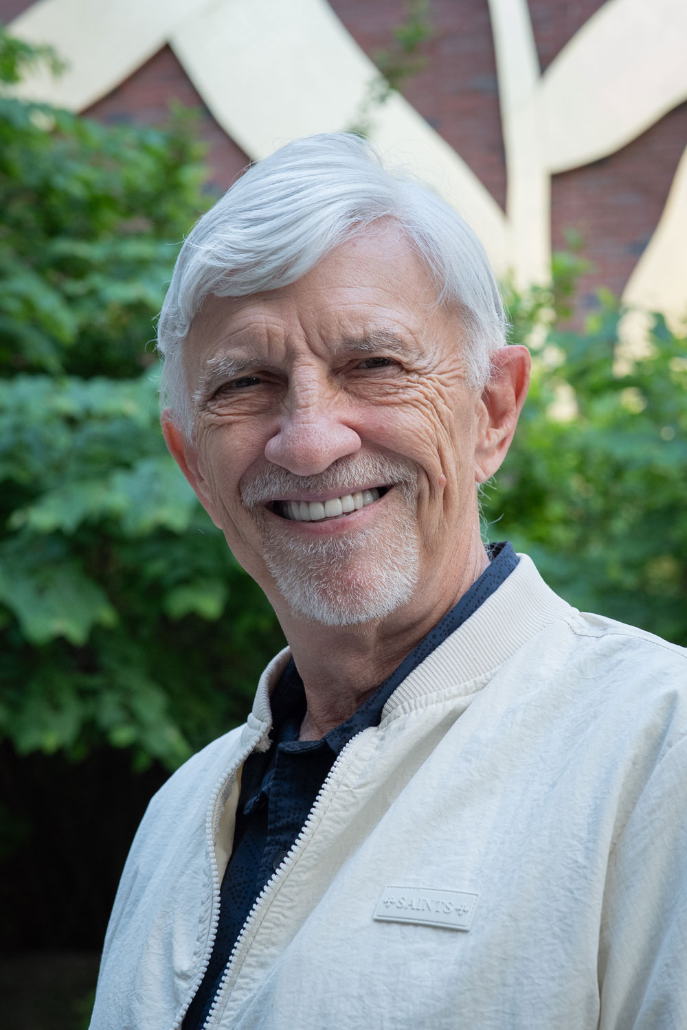 Chuck Gutenson smiles toward camera, posed in a white blazer against some foliage