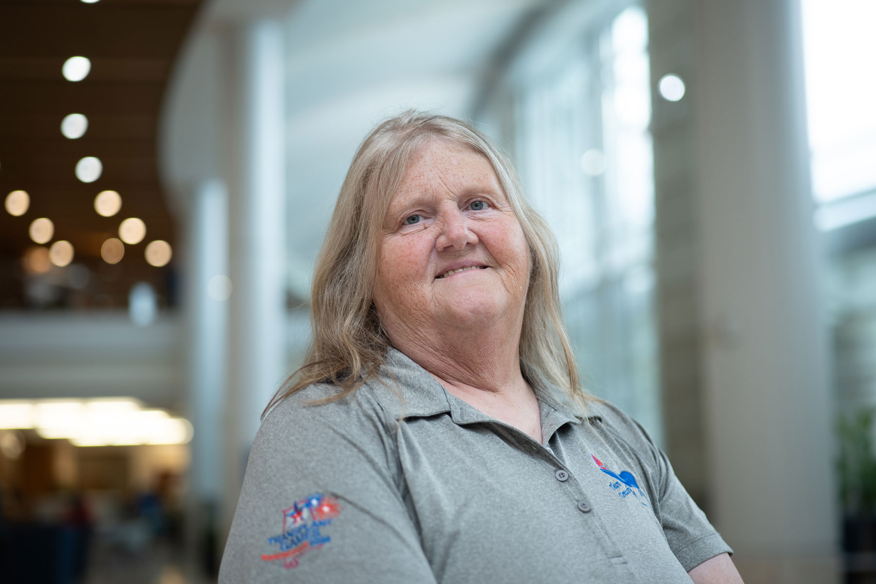 Linda Baxter poses for a portrait against the windows of the lobby in UK Albert B. Chandler Hospital. She's wearing a gray polo.
