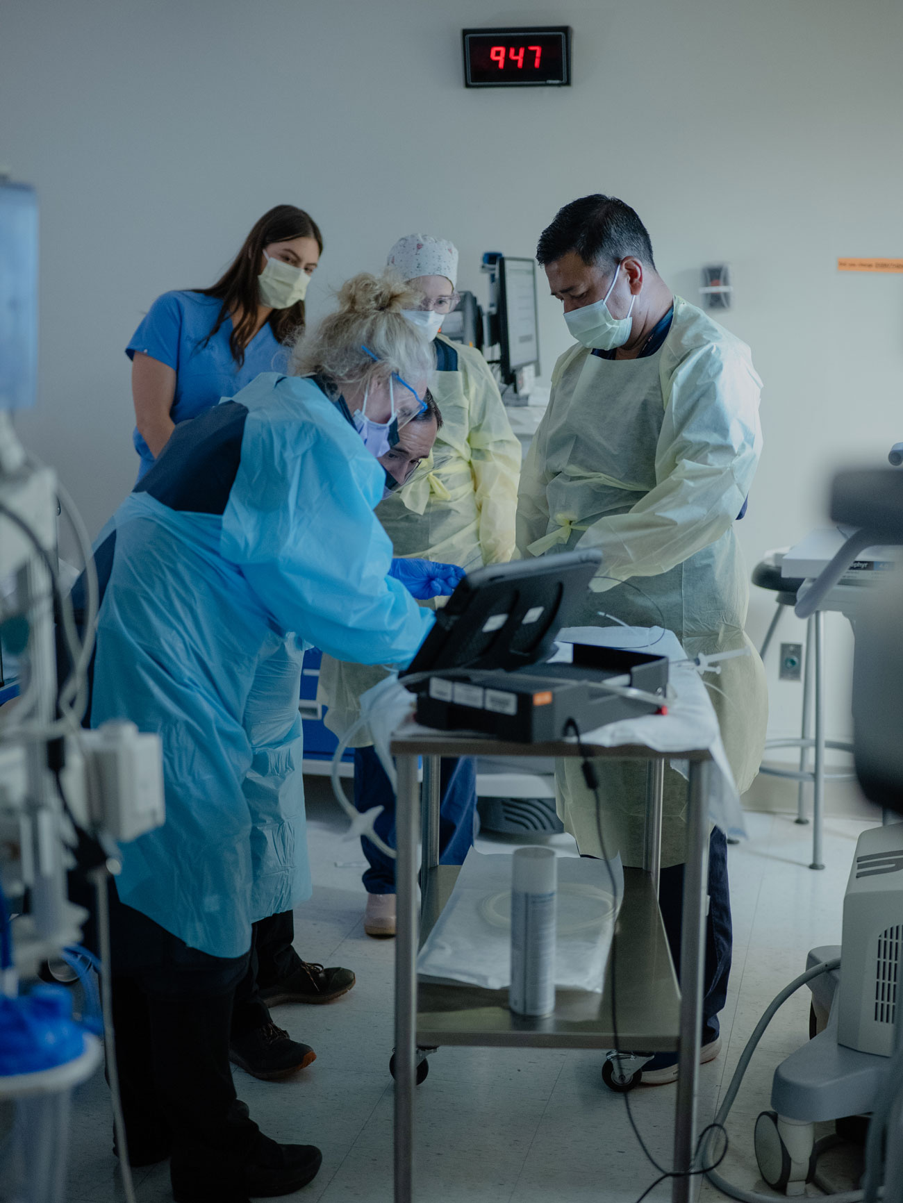 A group of medical professionals prepare for a procedure on a table. A clock reads 9:47 above them
