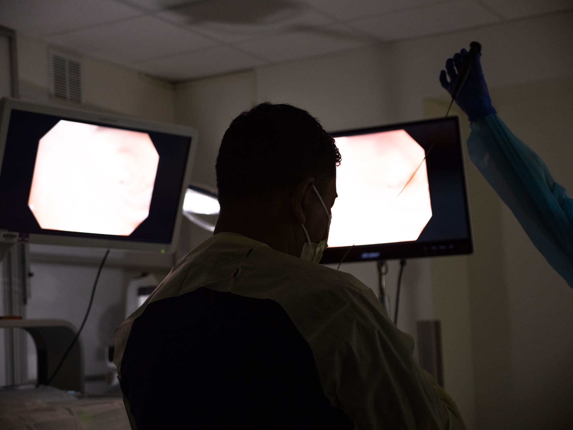 Dr. Maskey, shot from behind in a dark procedure room, with monitors displaying the inside of a patient's lungs. An unknown person's hand is holding a medical device.