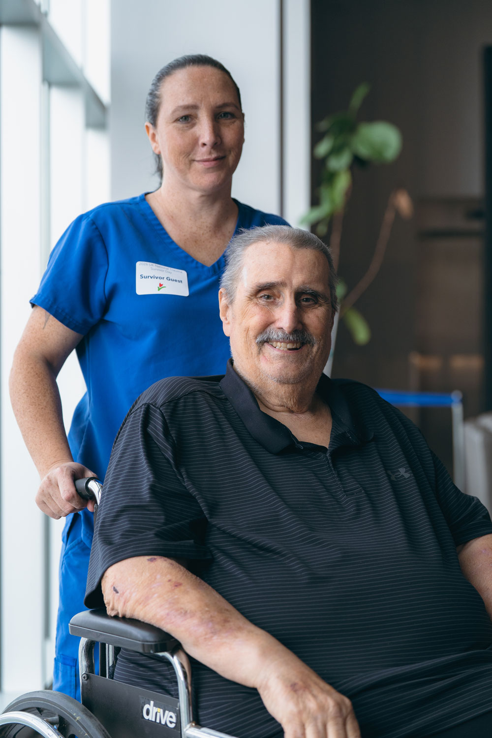 Jerry Gumm, seated in a wheelchair and smiling at camera, with an aide dressed in blue scrubs standing behind him