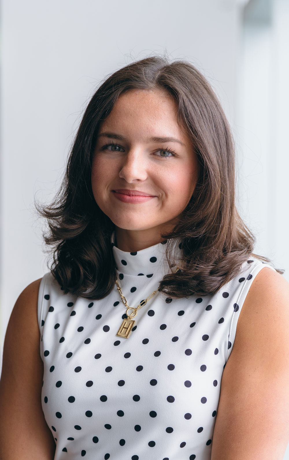Holly Harrison, wearing a gold "H" necklace and a white shirt with no sleeves and little flowers, smiles softly at camera