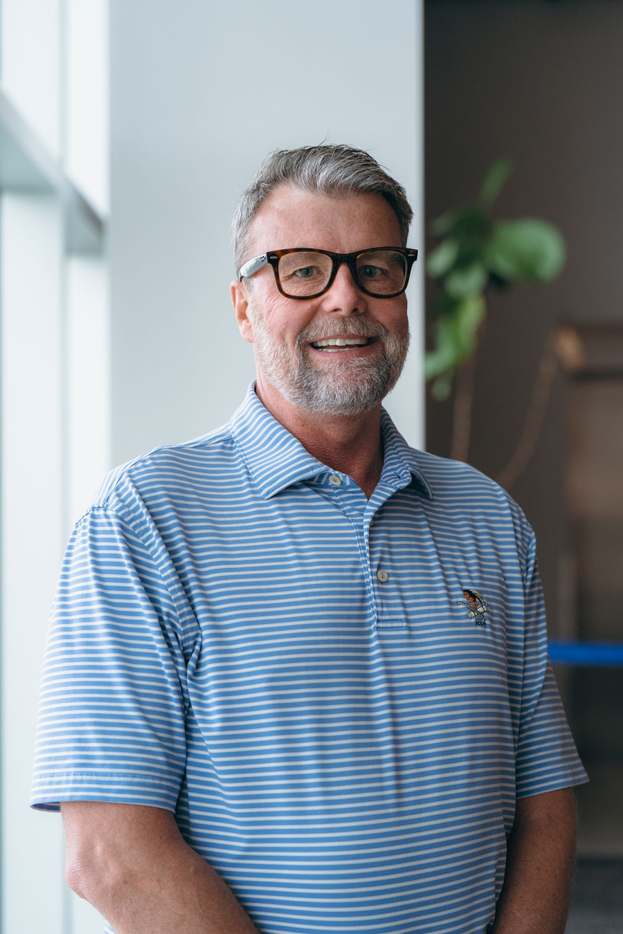 Michael Hudkins, smiling at camera, dressed in a blue striped polo and wearing thick glasses