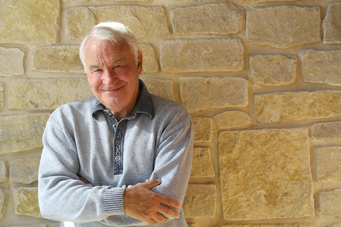 A smiling man wearing a gray sweater stands in front of a stone wall
