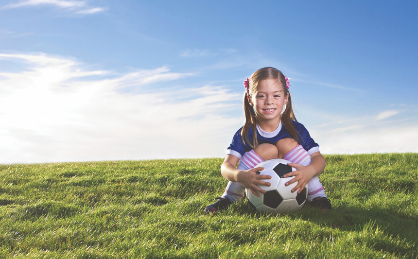 A young girl holding a soccer ball sits in a grassy field.