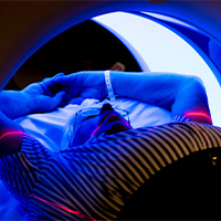 A person lies on a sliding table entering a large circular medical imaging scanner. The scene is illuminated with blue and pink lighting effects.