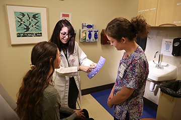 A female doctor wearing a white coat and a female nurse wearing floral scrubs meet with an adolescent female patient.