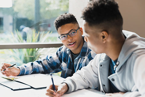 Two African American teenage boys study together at a table next to a window.