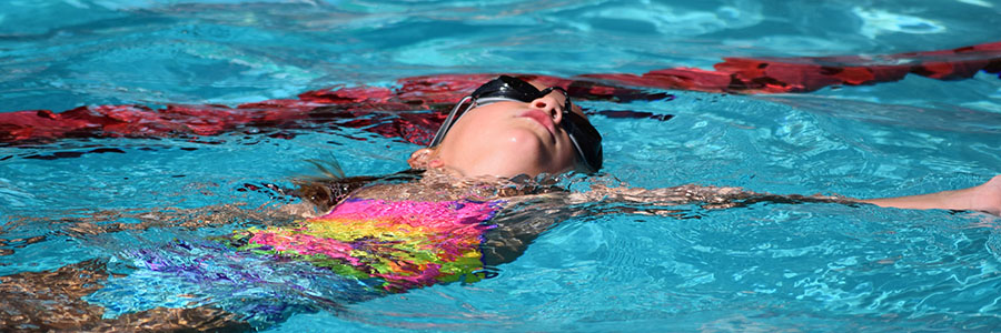 Teen girl wearing goggles and a tie-dye swimsuit doing the backstroke in a swimming pool