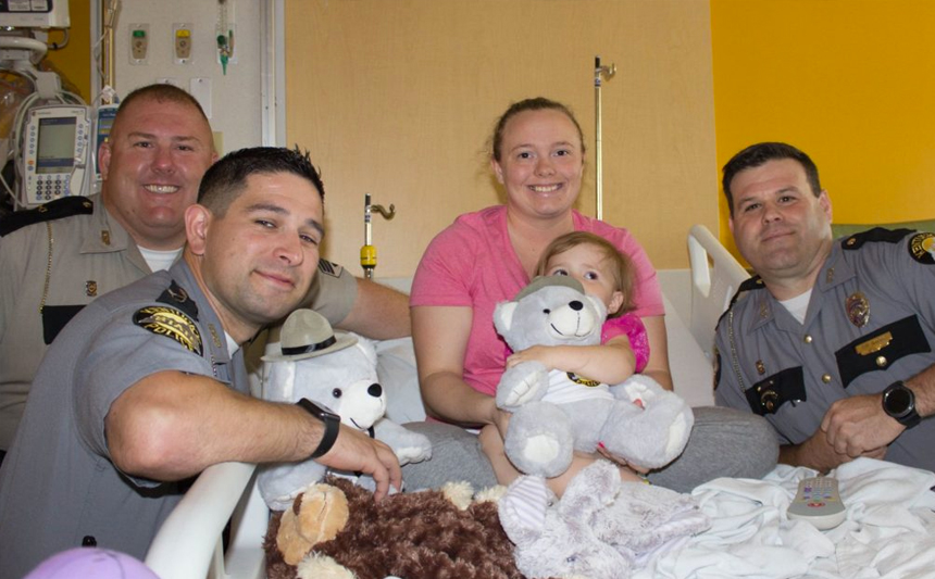 Three Kentucky State Troopers deliver a teddy bear to a young girl patient.