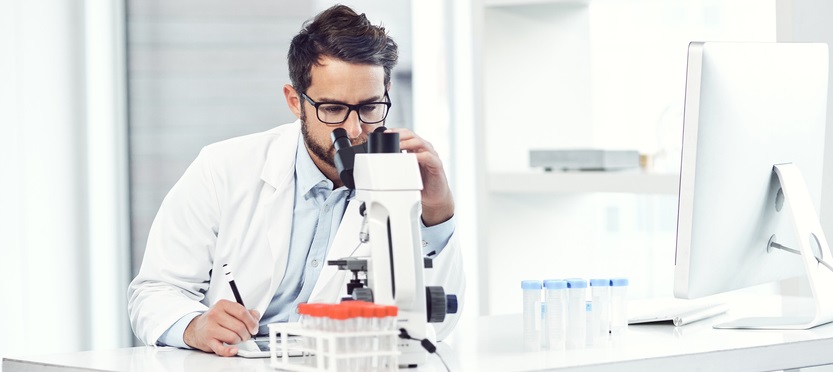 Young male scientist making notes while looking trough a microscope 