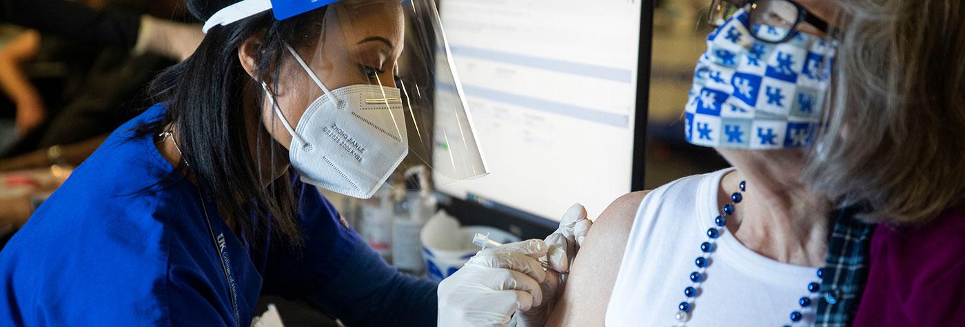 A nurse administers a COVID-19 vaccine to a woman wearing glasses, a University of Kentucky-branded face mask, a white sleeveless blouse, and a blue necklace.