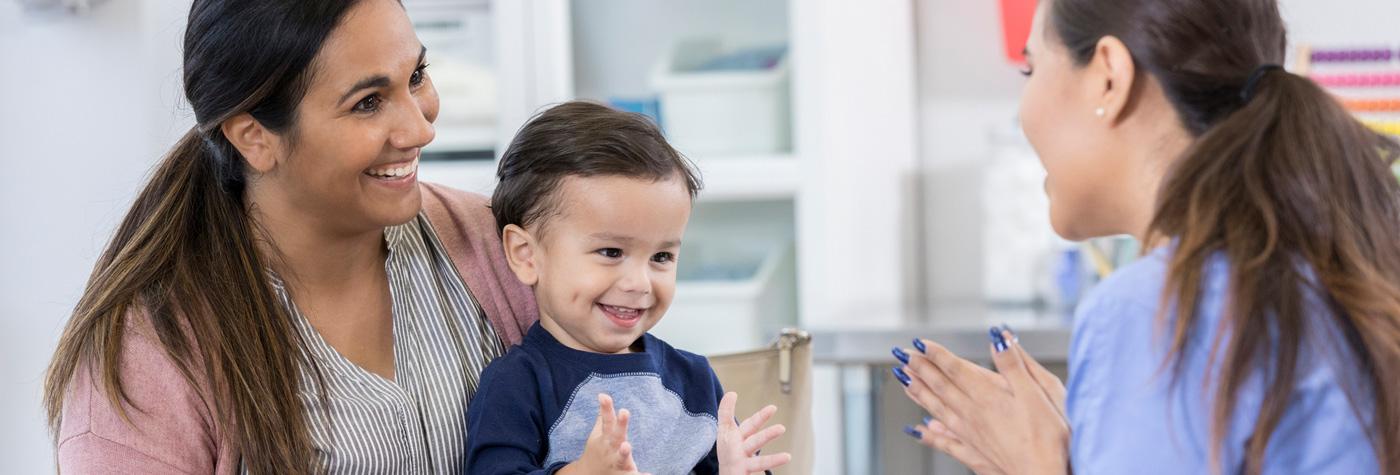 Female provider wearing blue scrubs talks to a smiling toddler while he sits on his mother's lap. 