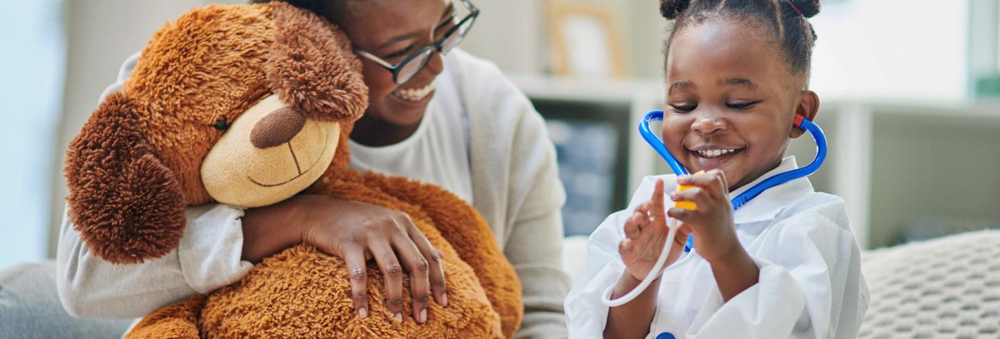 A young African-American girl uses a toy stethoscope on a stuffed animal while her mother, sitting next to her, laughs.