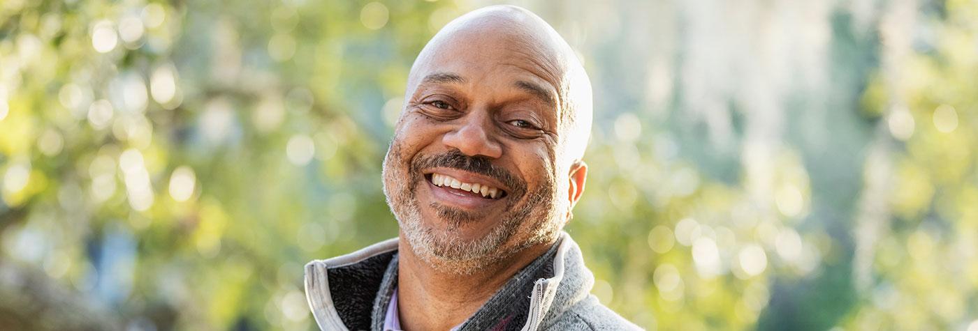 Middle aged African American man standing outside in front of trees smiles at the camera. 