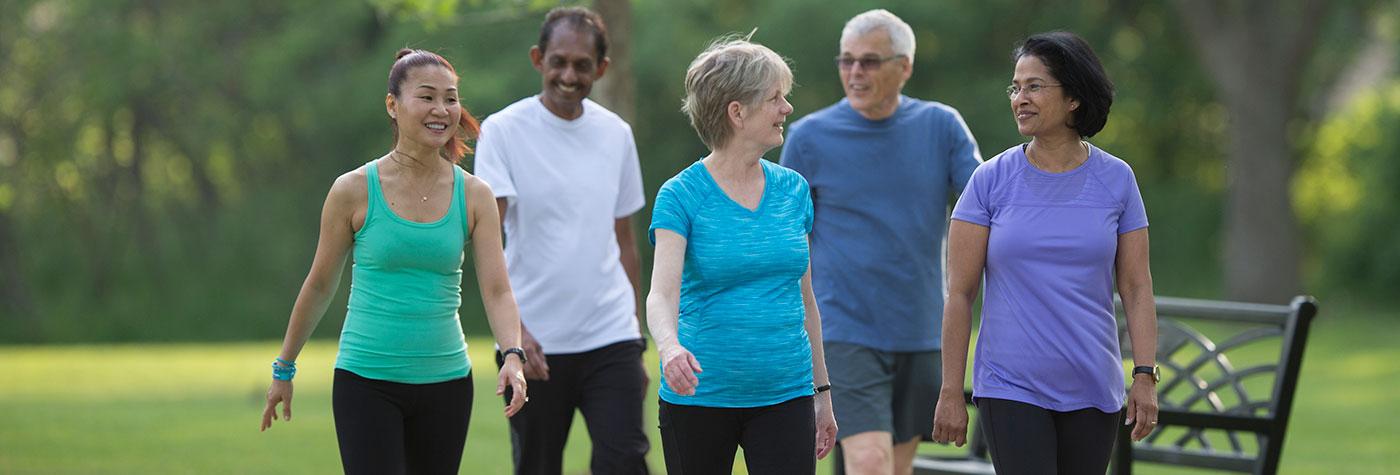 A diverse group of adults wearing athletic attire walks and talks in a park. 
