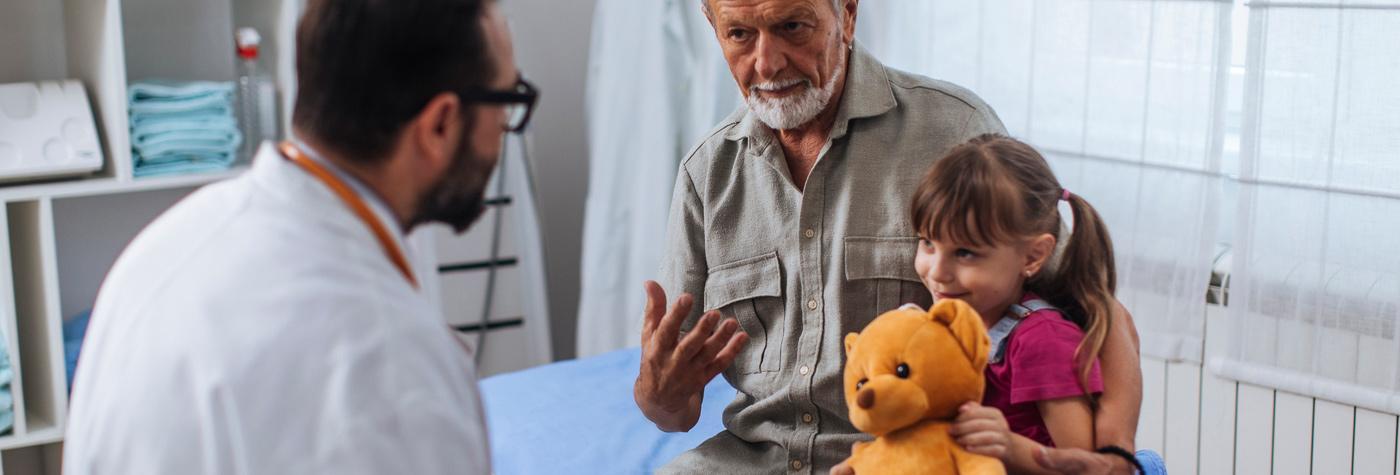 A doctor speaks with a young girl, who's holding a teddy bear, and the family member who accompanied her to the appointment.