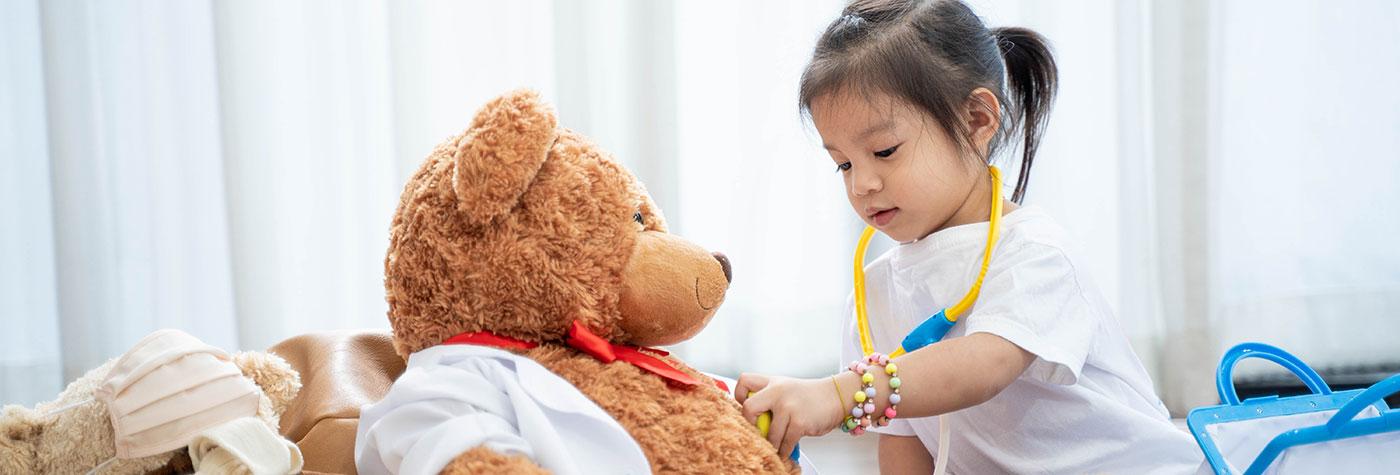 A preschool-aged girl uses a toy stethoscope and pretends to listen to a large teddy bear's heartbeat.