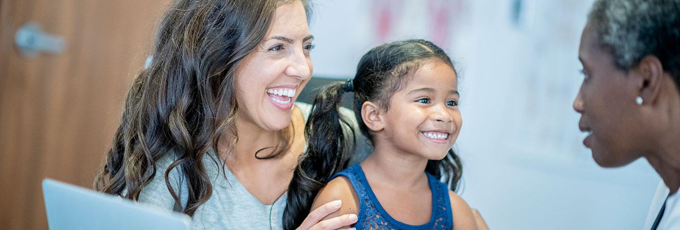 A girl of hispanic ethnicity smiles while she sits in her mother's lap. Her mother is smiling too, as she listens to what their doctor has to say. There is an open laptop out on the table.