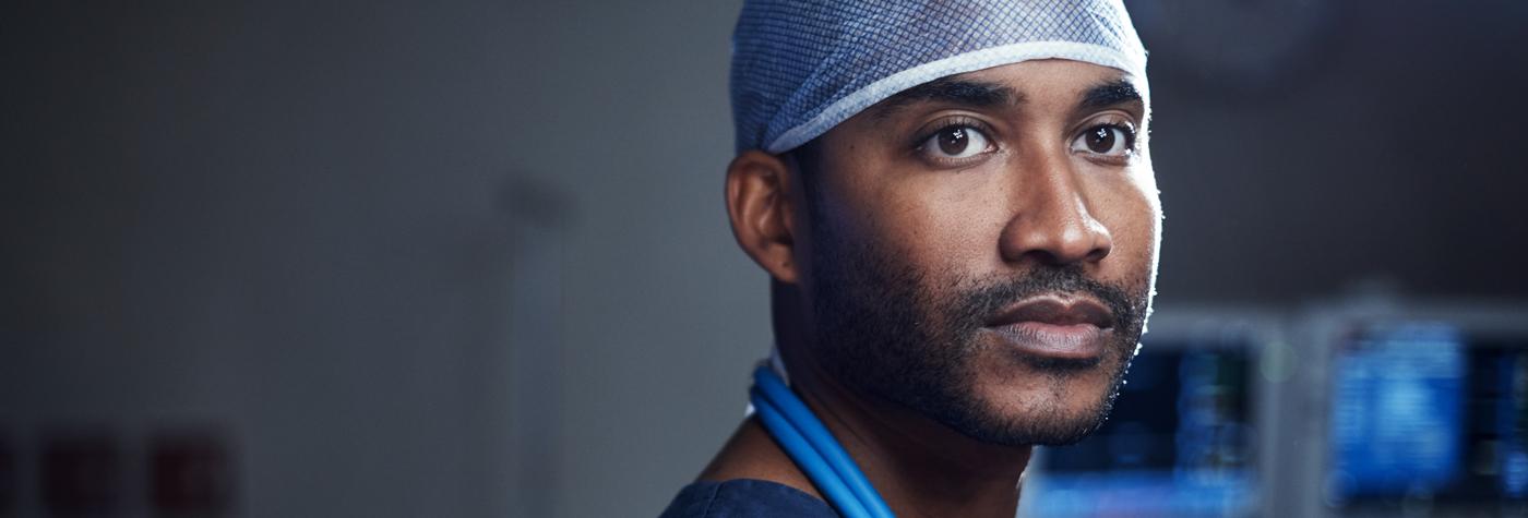 African American man in blue scrubs and surgical cap looks toward the camera with a thoughtful expression on his face. 