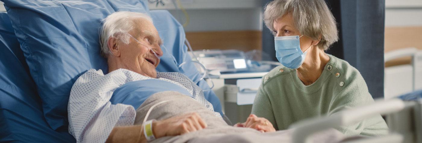 An older woman wearing a mask talks with her partner, an older man, who is sitting up in a hospital bed and wearing a gown and nasal cannula. 