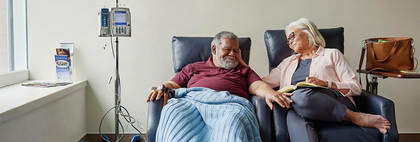 An older African-American man sits in a comfortable chair with a light blue blanket over his lap while receiving medication via an IV pump. His wife sits in a chair next to him, with her hand gently touching his face.