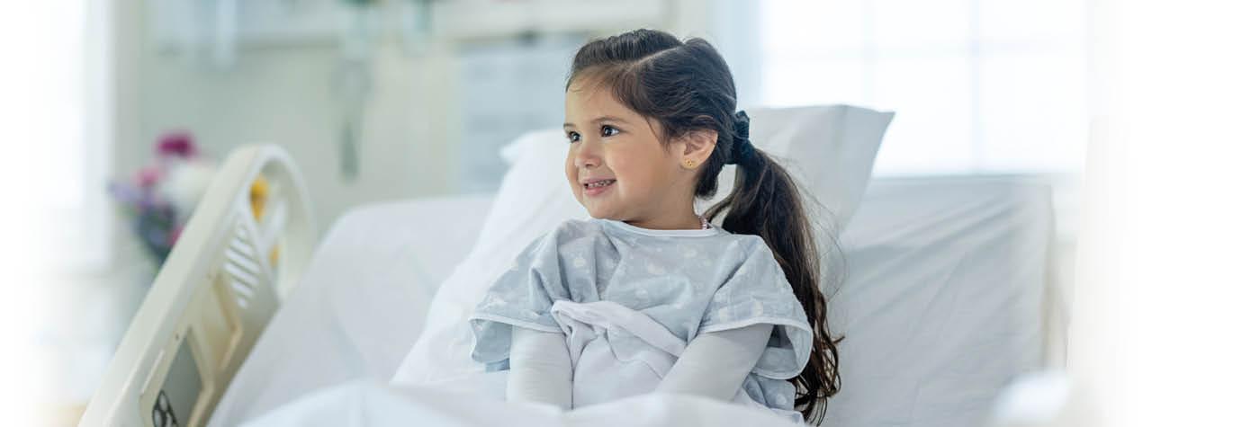 A smiling young girl wears a hospital gown while sitting propped up by a pillow in a hospital bed.