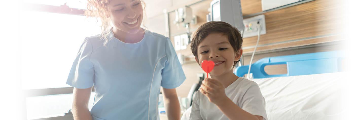 A little boy sits in a hospital bed and smiles while holding a red lollipop. A nurse wearing a light blue scrub top looks on next to him. She is also smiling. 